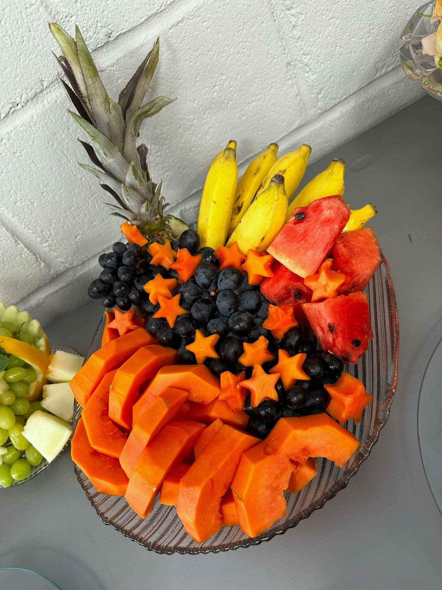 Travessa com mamão fatiado, uvas pretas, frutas cortadas em estrela, bananas, melancia e coroa de abacaxi.