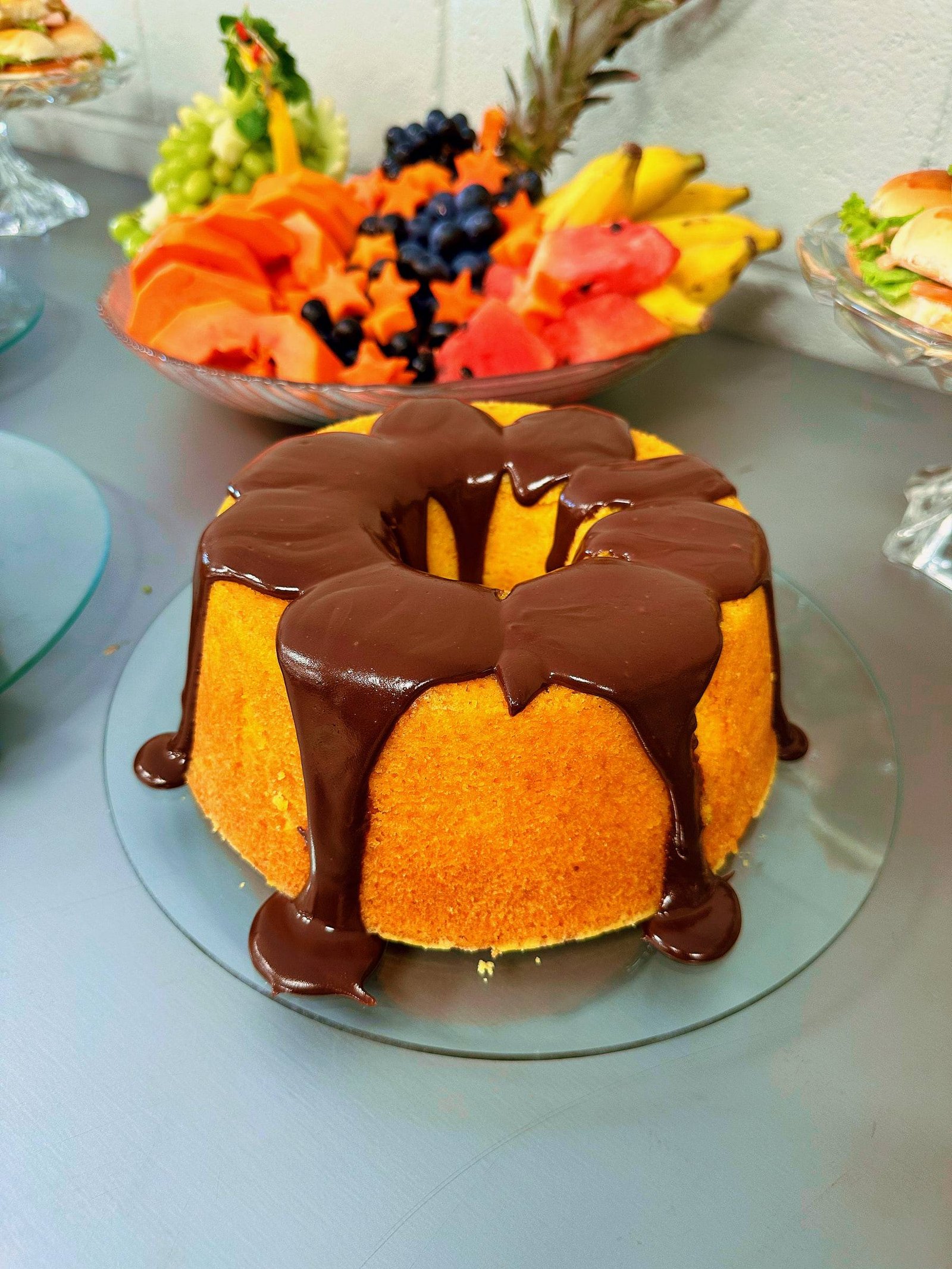 Bolo com calda de chocolate escorrendo, em prato de vidro, com travessa de frutas ao fundo.