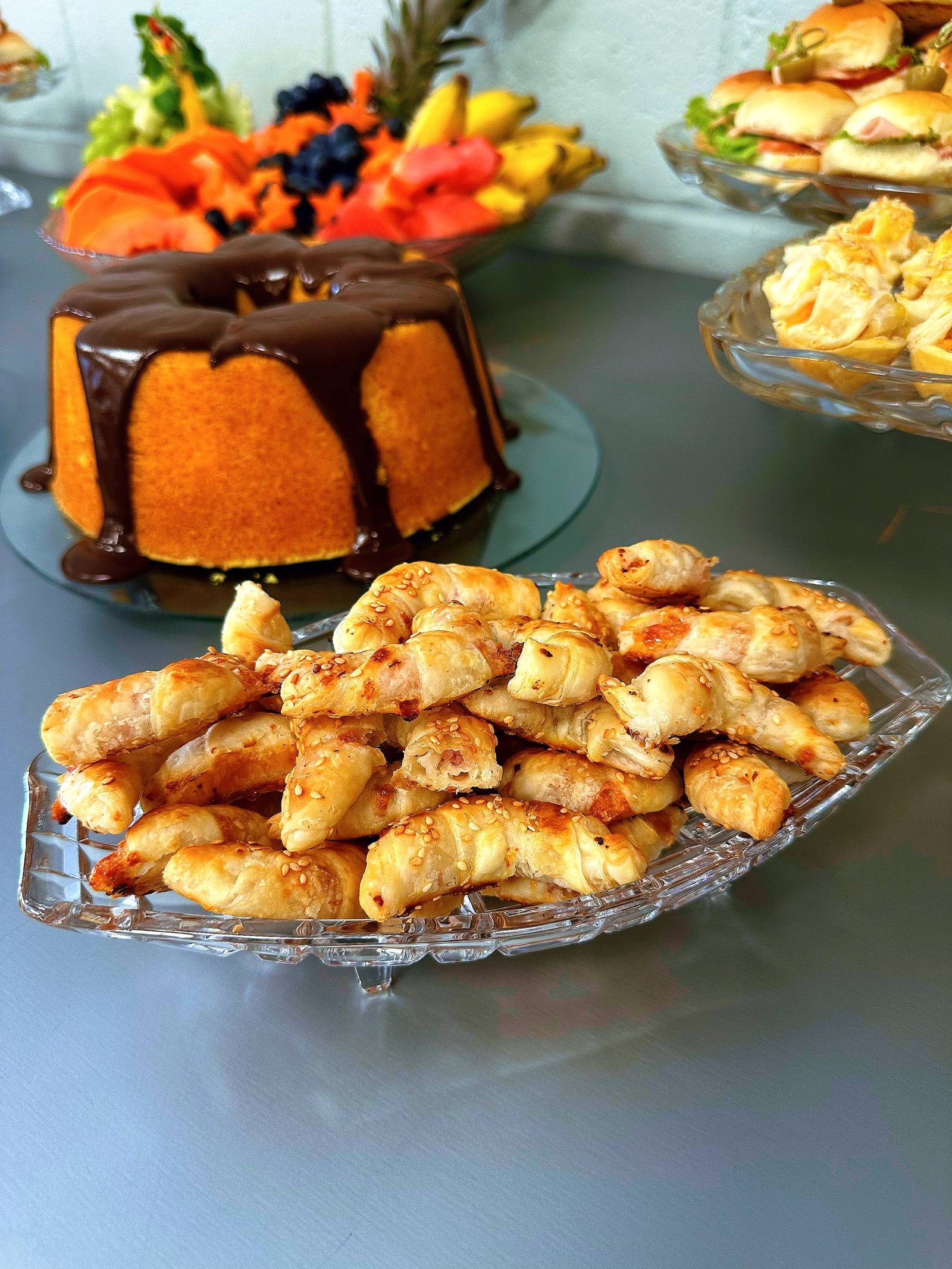 Travessa de cristal com folhados, com bolo com calda de chocolate e frutas ao fundo.