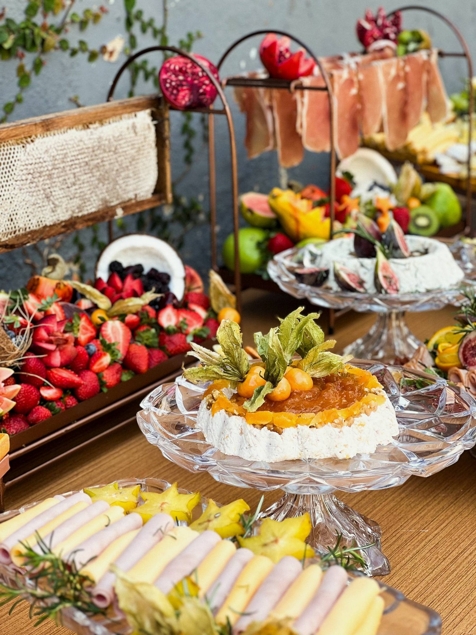 Mesa com morangos, frutas tropicais, pastas decoradas, frios enrolados e presunto em suporte de ferro, ao lado de favo de mel.