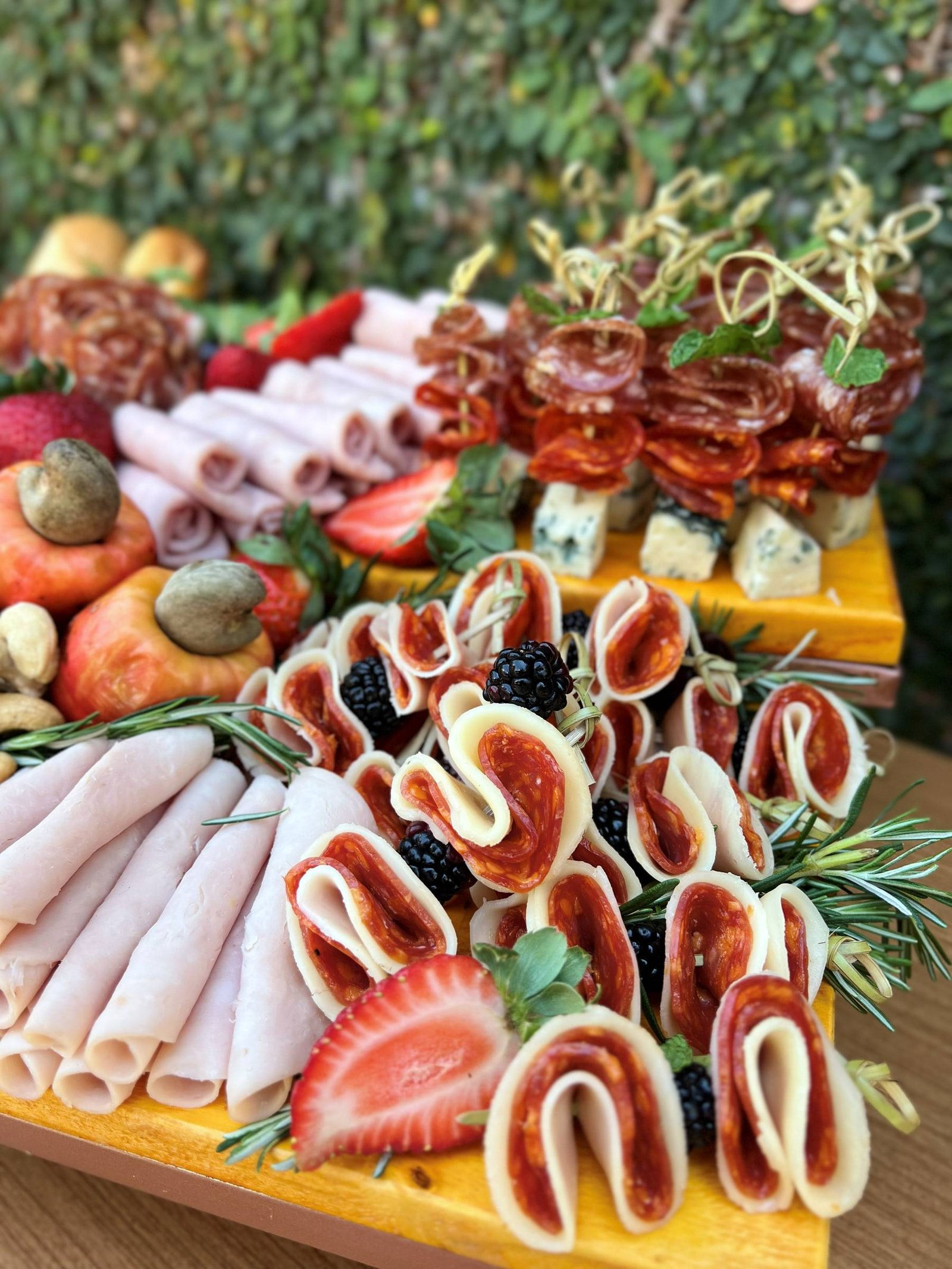 Tábua com frios enrolados, salame, morangos, amoras e queijo, com espetinhos de salame ao fundo.